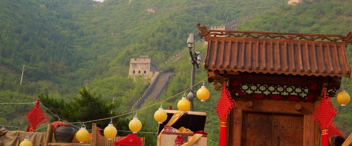 A traditional Chinese building with a tiled roof and red decorations, set against a backdrop of a large, ancient wall and lush green hills.