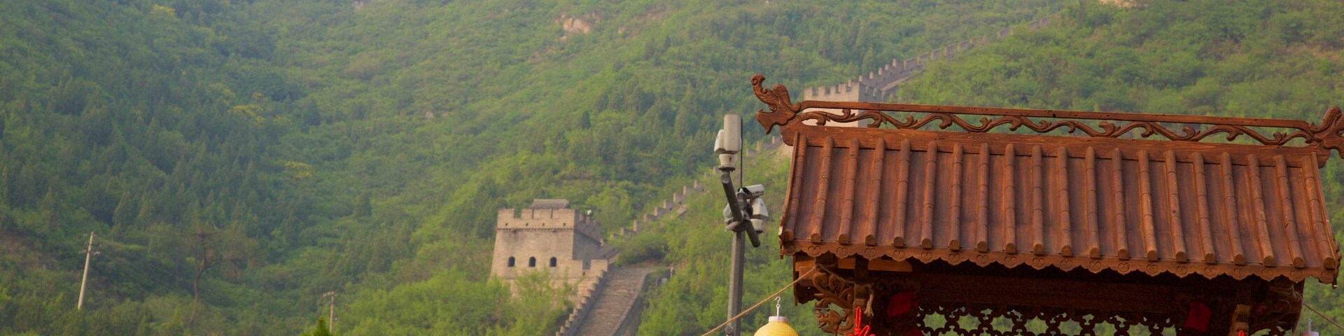 A traditional Chinese building with a tiled roof and red decorations, set against a backdrop of a large, ancient wall and lush green hills.
