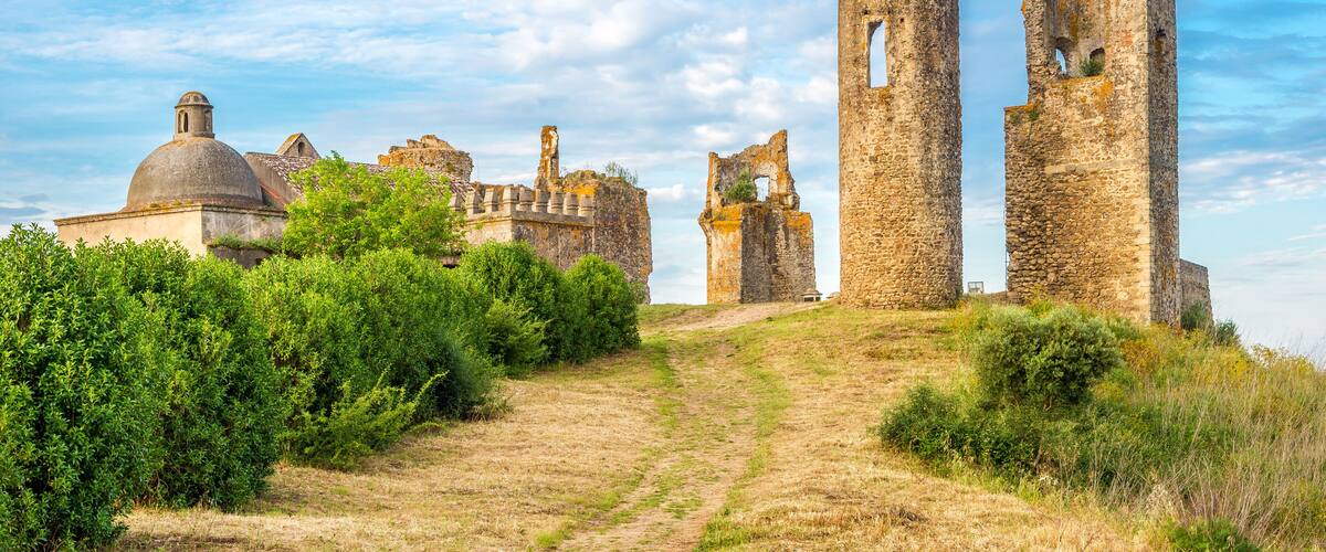 A historic stone castle with ruins, including a large tower and a dome, surrounded by greenery and a clear sky.
