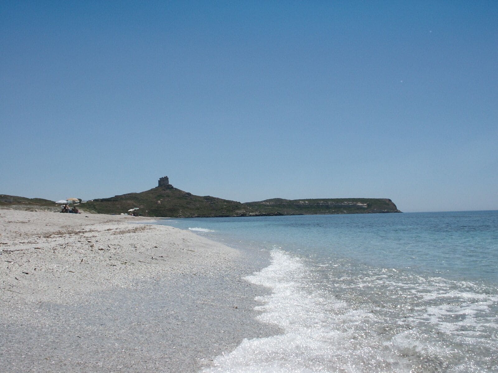 A sandy beach with gentle waves, a small hill with a structure on top, and a distant cliff.