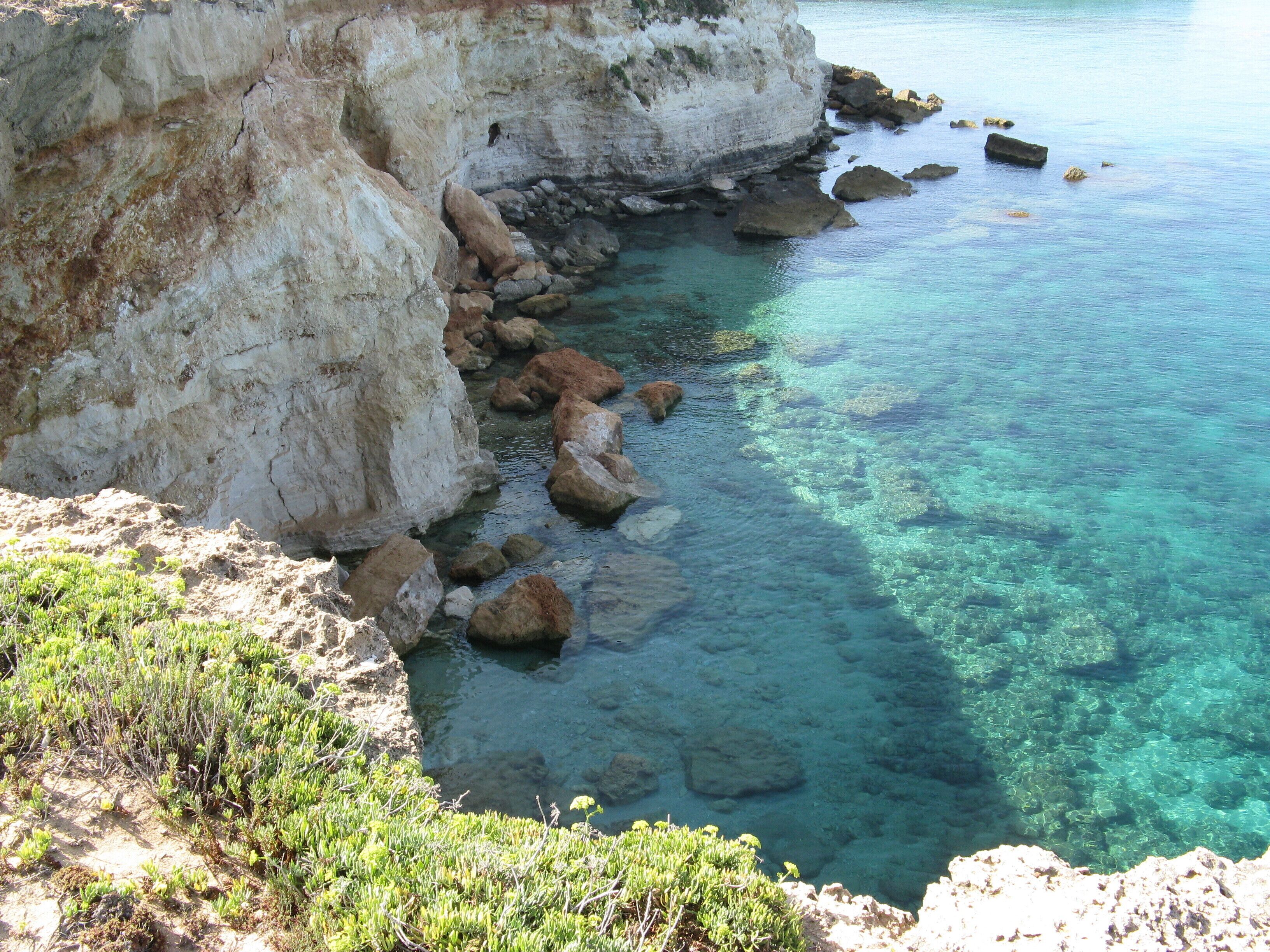 A coastal cliff with clear turquoise water and scattered rocks.