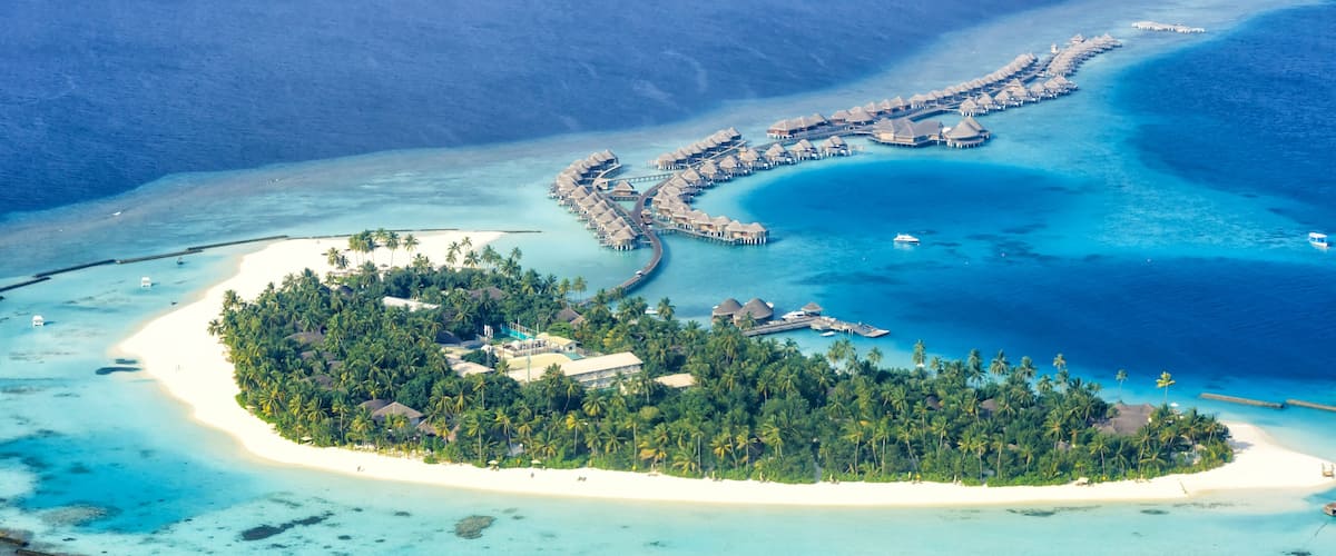 An aerial view of a tropical island with a white sandy beach, dense palm trees, and overwater bungalows connected by a dock.