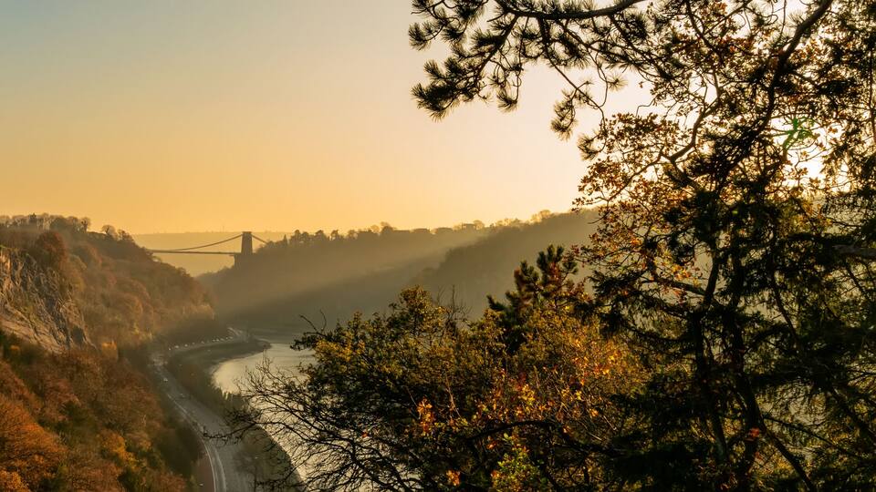 A river valley with autumn foliage, a bridge, and a mountainous backdrop.