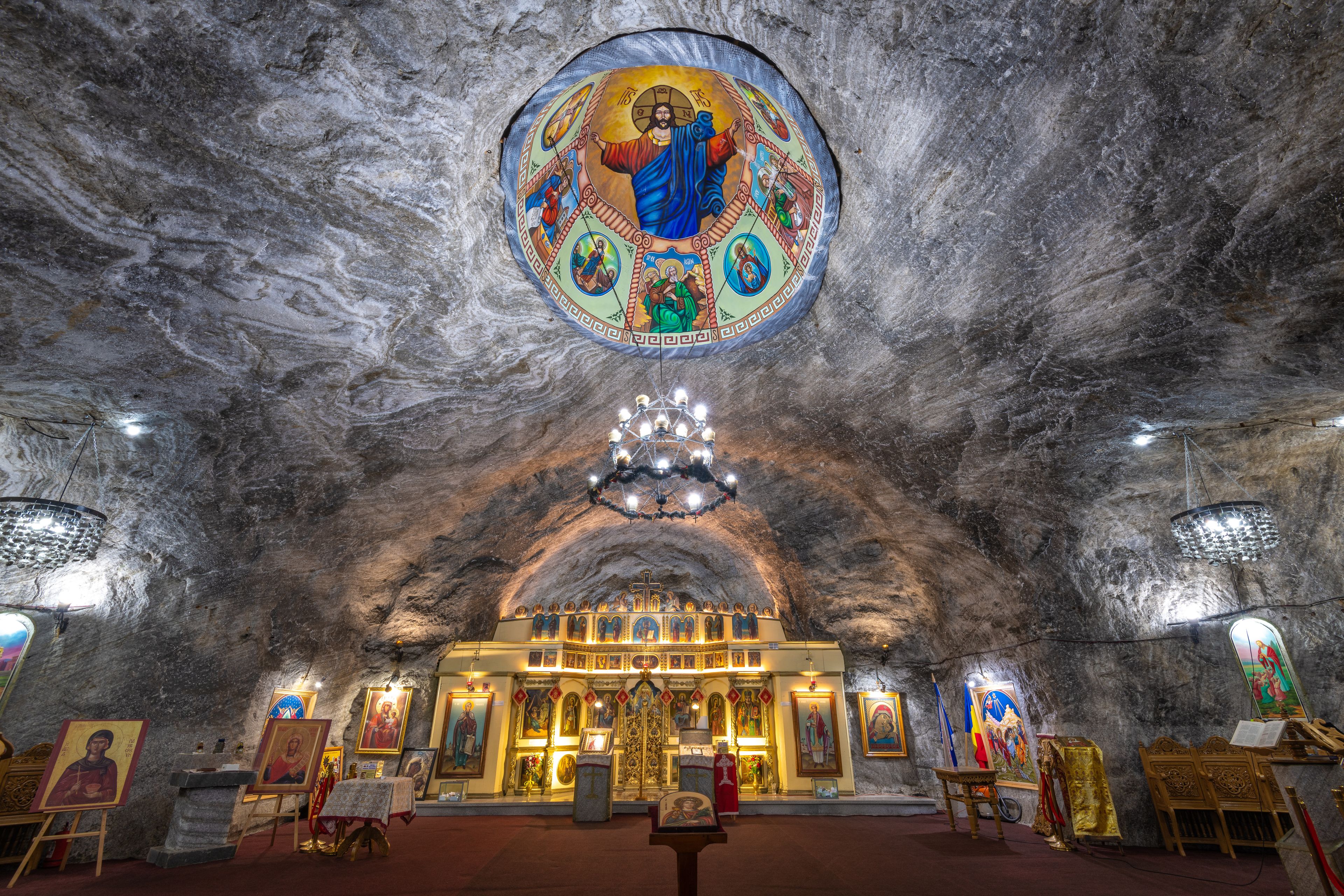 An interior of a cave church with a large circular icon above the altar, stone walls, and a chandelier.