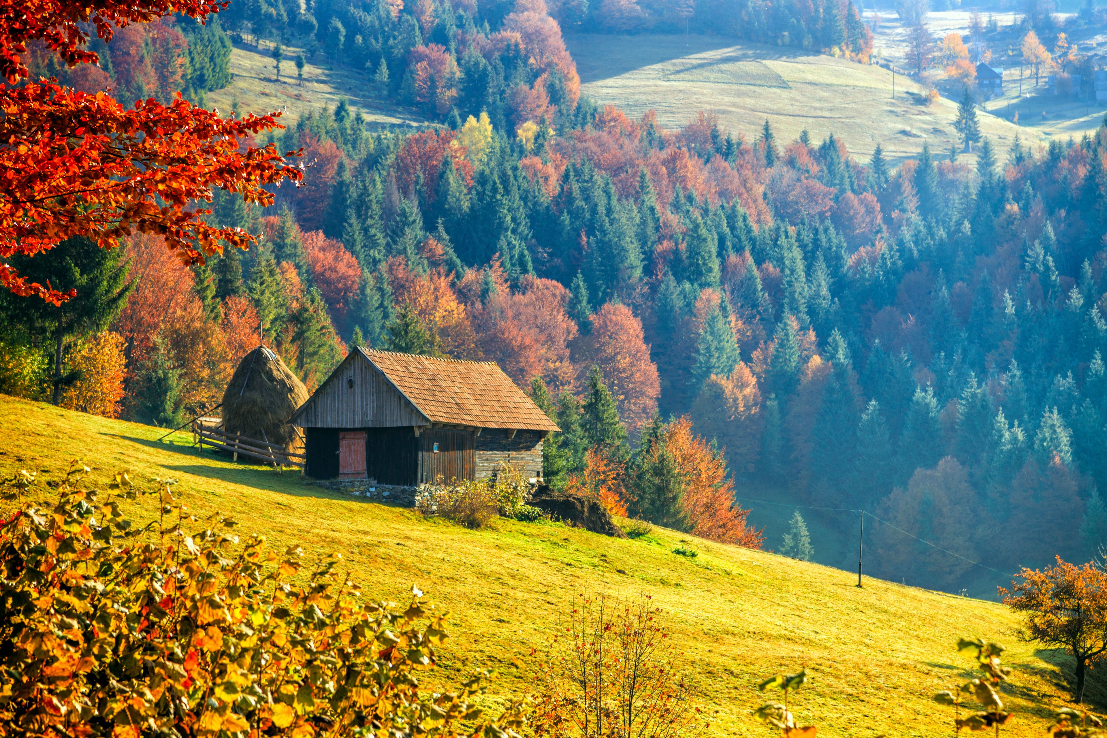 A rural landscape with a wooden barn on a hillside, surrounded by autumn-colored trees and rolling fields.