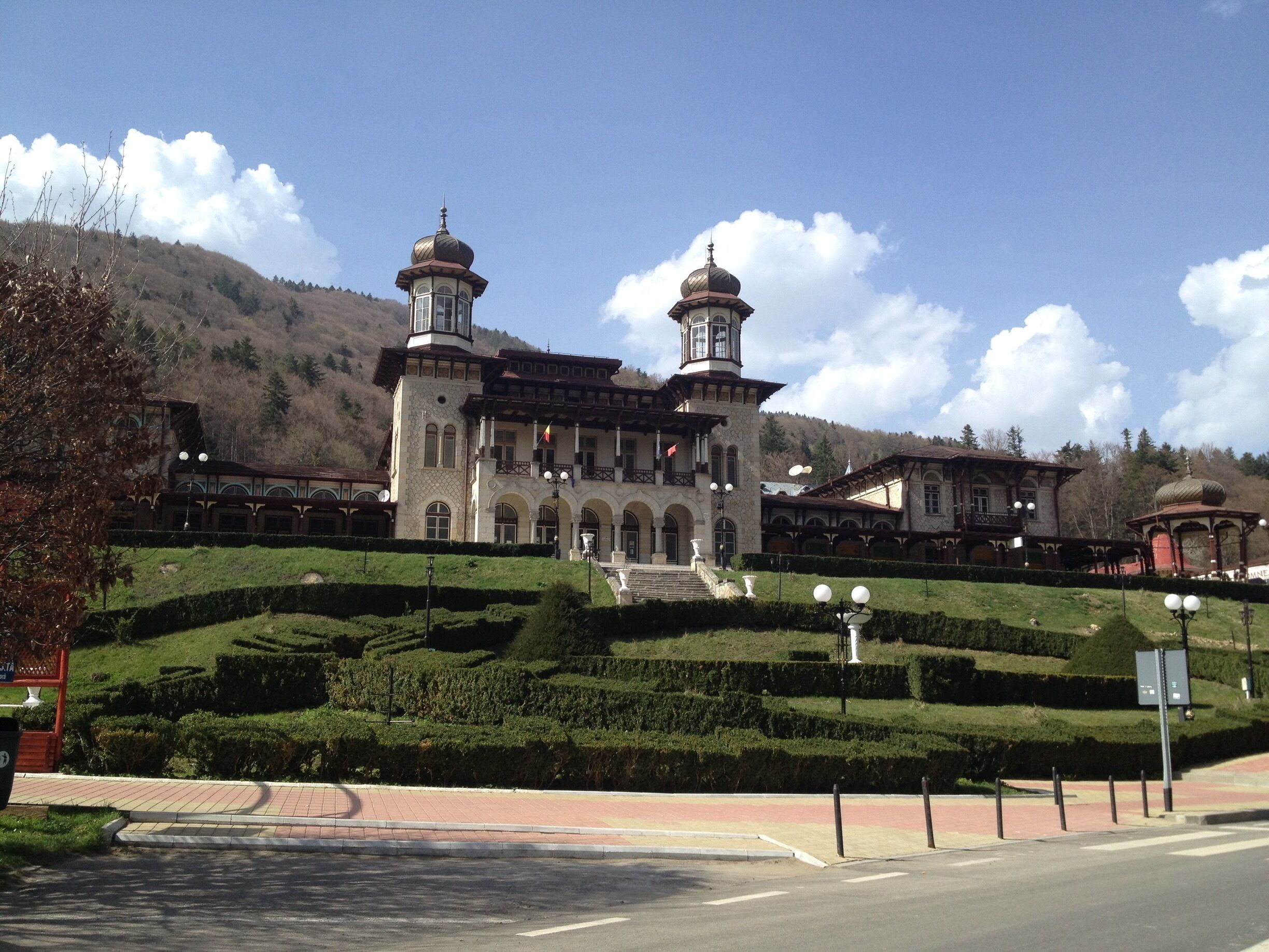 A historic building with two towers, surrounded by well-manicured gardens and hills.