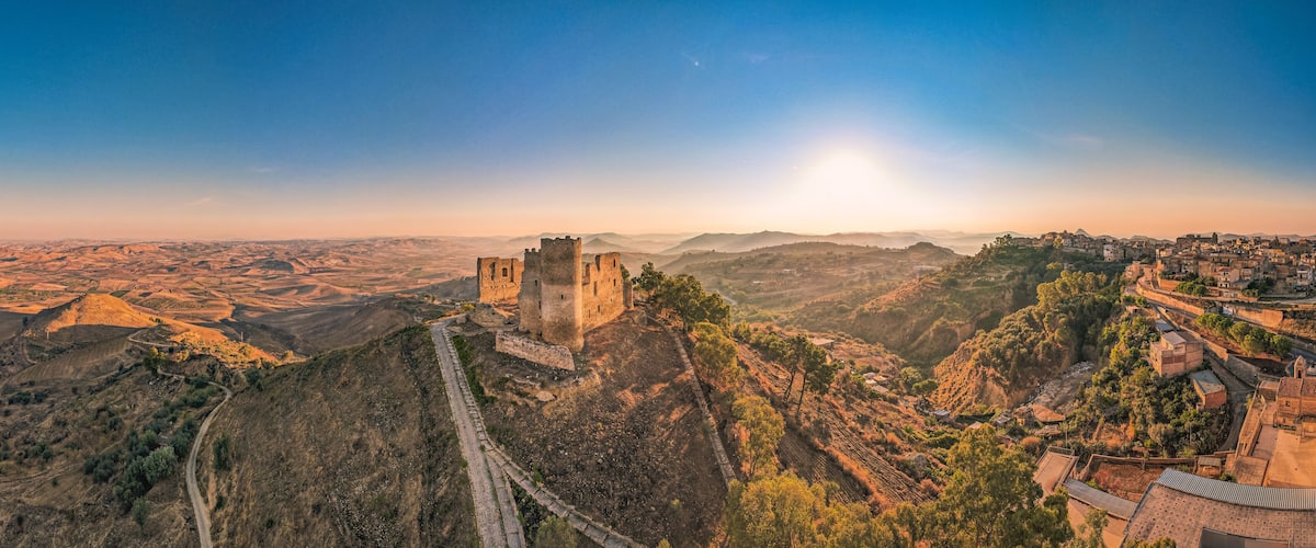 A panoramic view of a mountainous landscape with a prominent castle on a hill, surrounded by terraced fields and a small village.