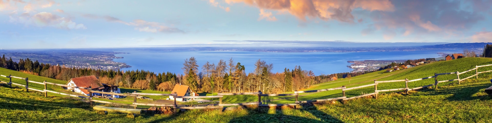 A panoramic view of a coastal landscape with a wooden fence, houses, and a distant body of water.