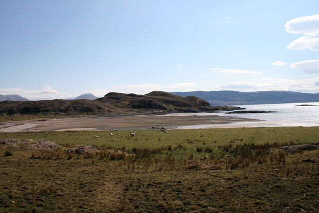 Un paisaje costero con campos verdes, una playa y montañas lejanas bajo un cielo despejado.