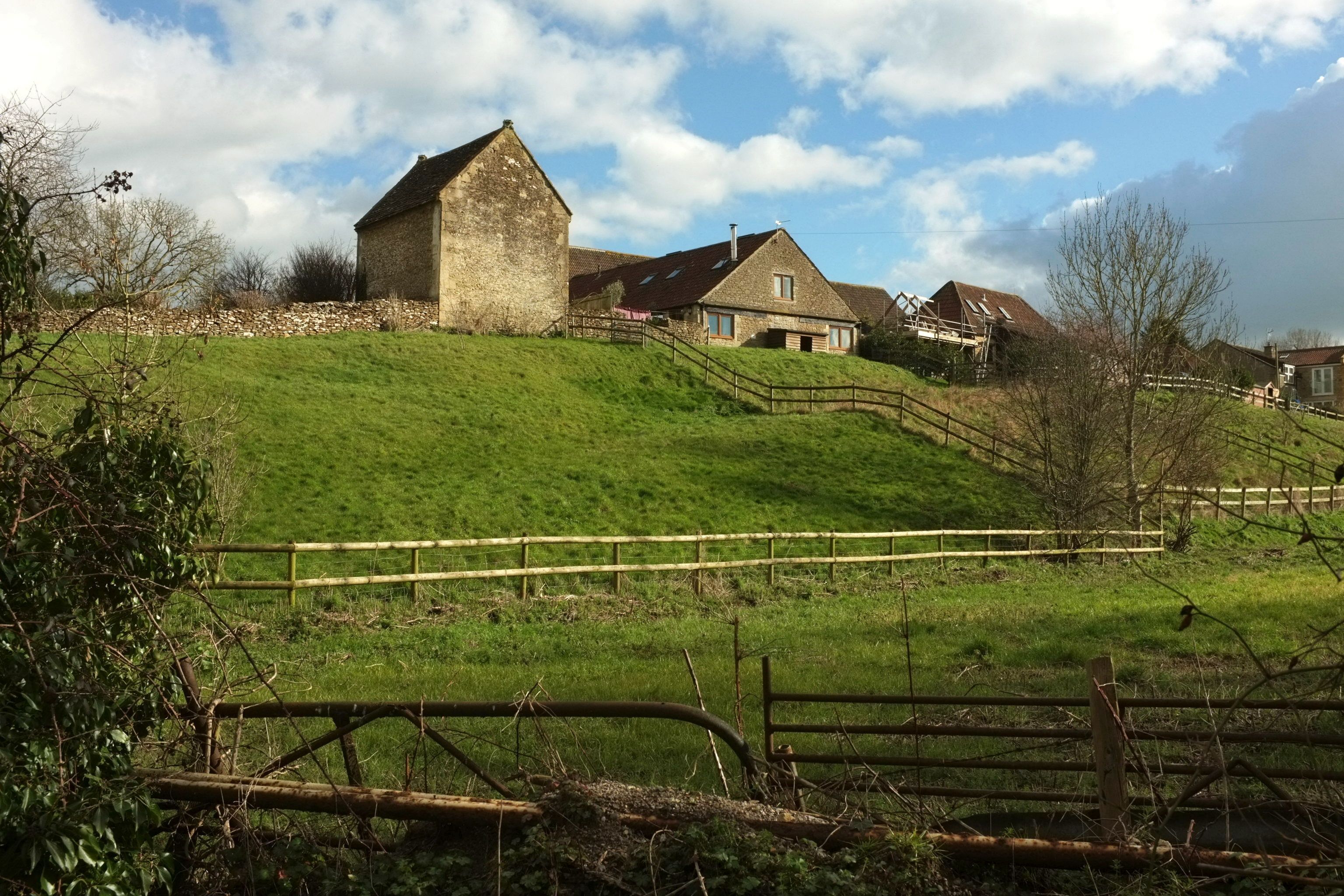 A rural landscape with a stone house, wooden fences, and a grassy hill.