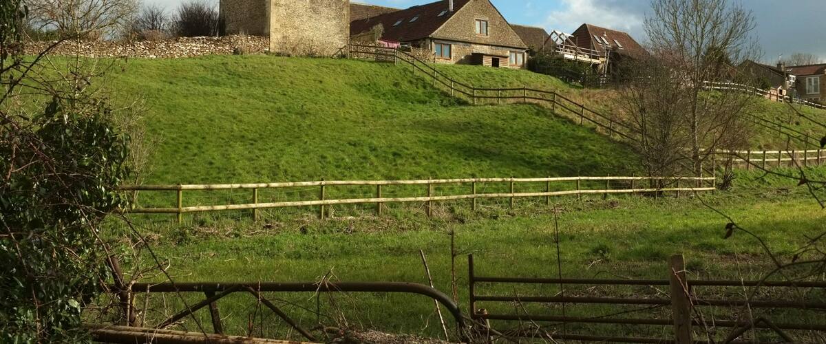A rural landscape with a stone house, wooden fences, and a grassy hill.