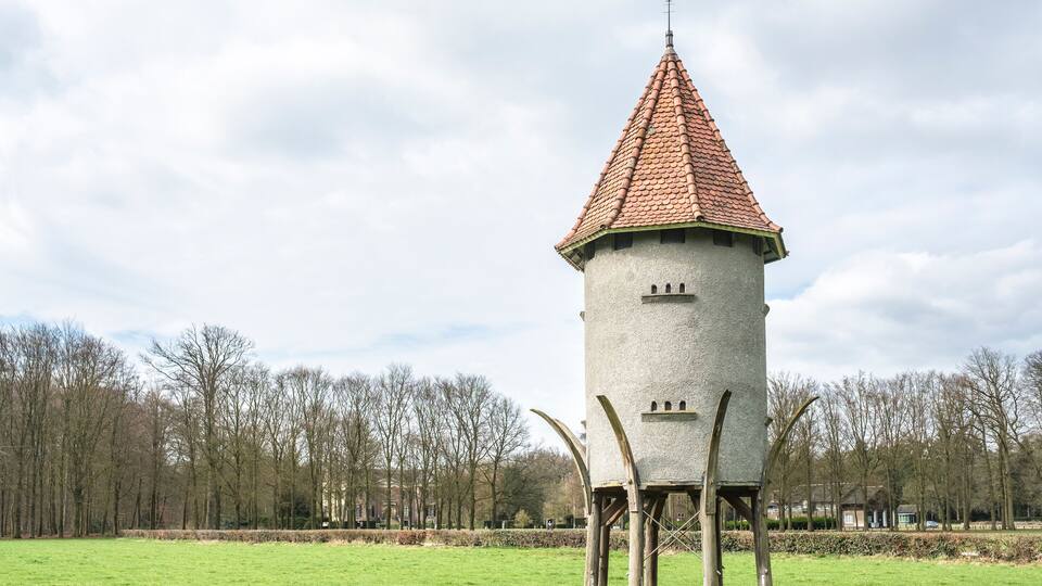 A tower with a conical roof and a flag atop, surrounded by a grassy field and trees.