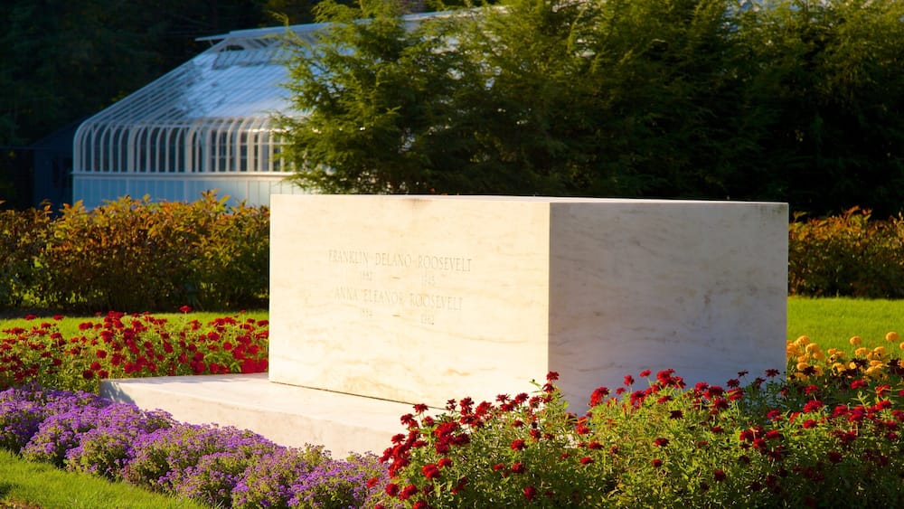 Un monument de marbre orné d’inscriptions, entouré d’un jardin où poussent des fleurs rouges et violettes, avec une serre en arrière-plan.