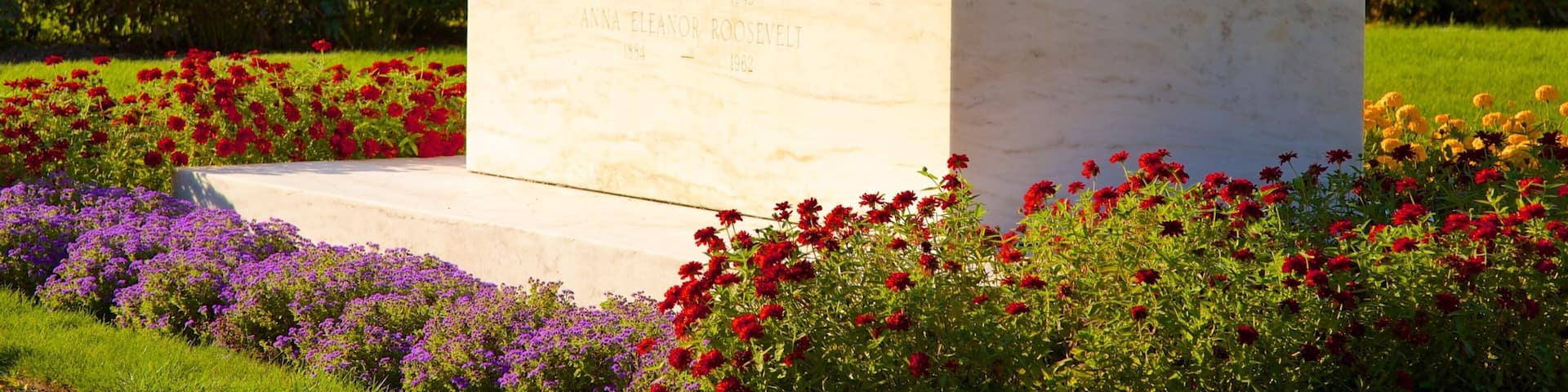 A marble monument with inscriptions, surrounded by a garden with red and purple flowers, and a greenhouse in the background.