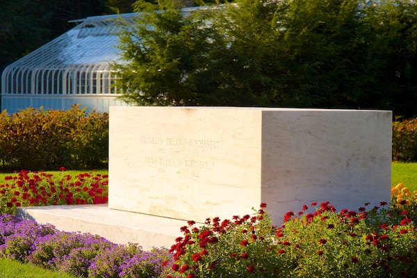 Un monument de marbre orné d’inscriptions, entouré d’un jardin où poussent des fleurs rouges et violettes, avec une serre en arrière-plan.