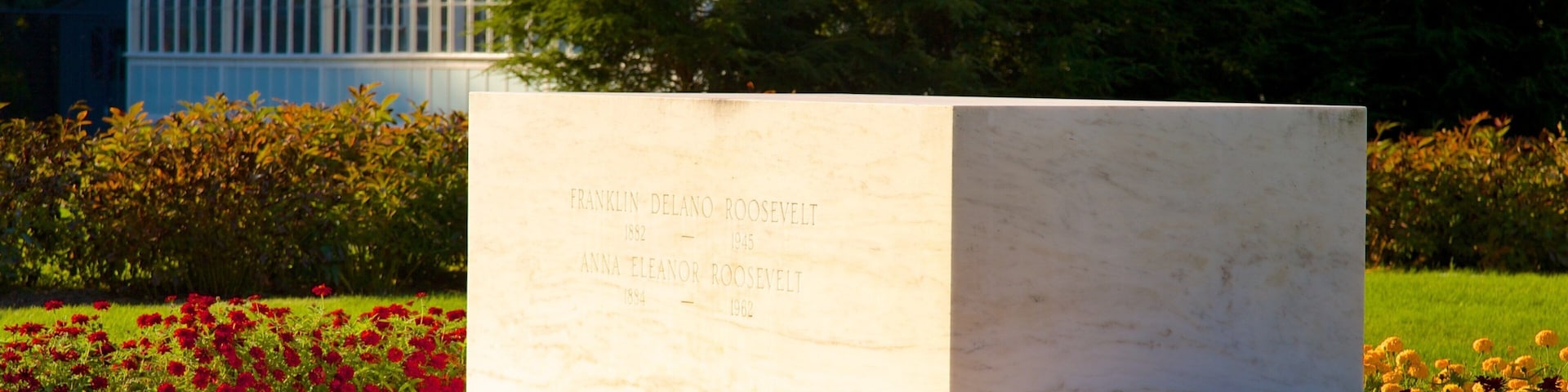 Un monumento de mármol con inscripciones, rodeado por un jardín con flores rojas y púrpuras, y un invernadero al fondo.