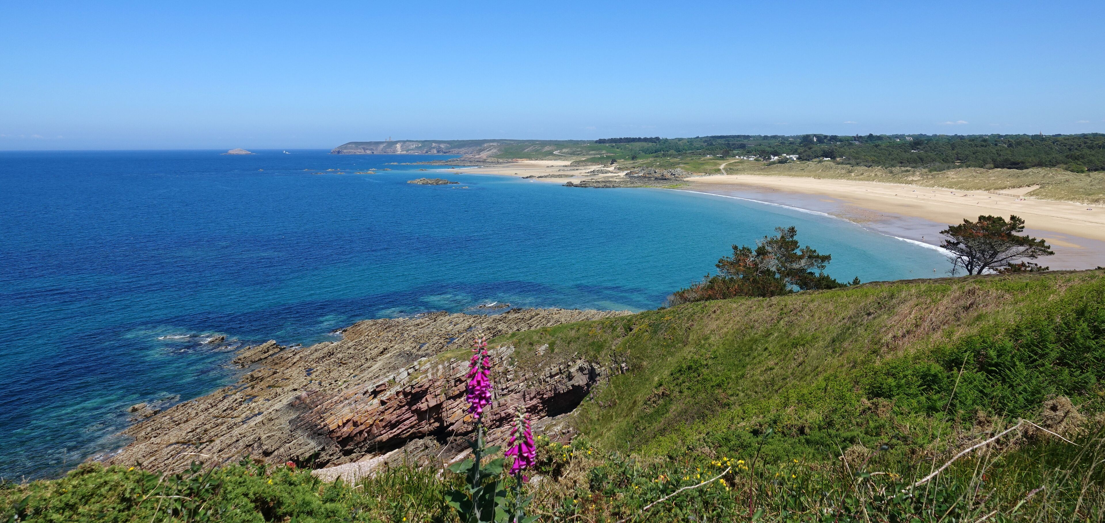 A coastal landscape with a sandy beach, clear blue waters, rocky outcrops, and green vegetation.