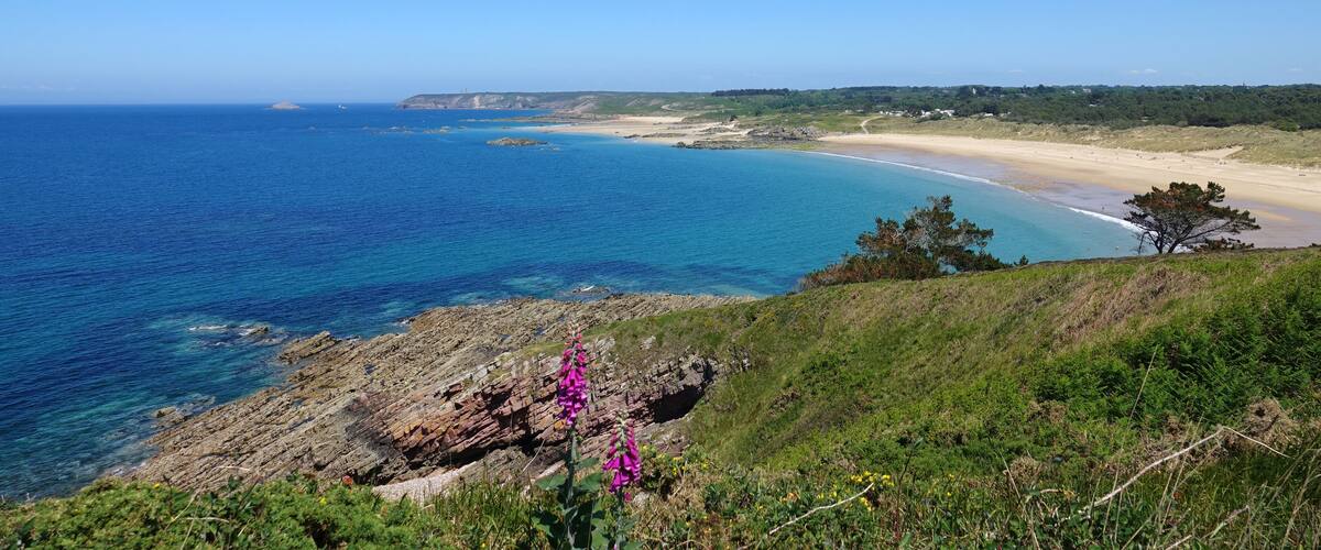 A coastal landscape with a sandy beach, clear blue waters, rocky outcrops, and green vegetation.