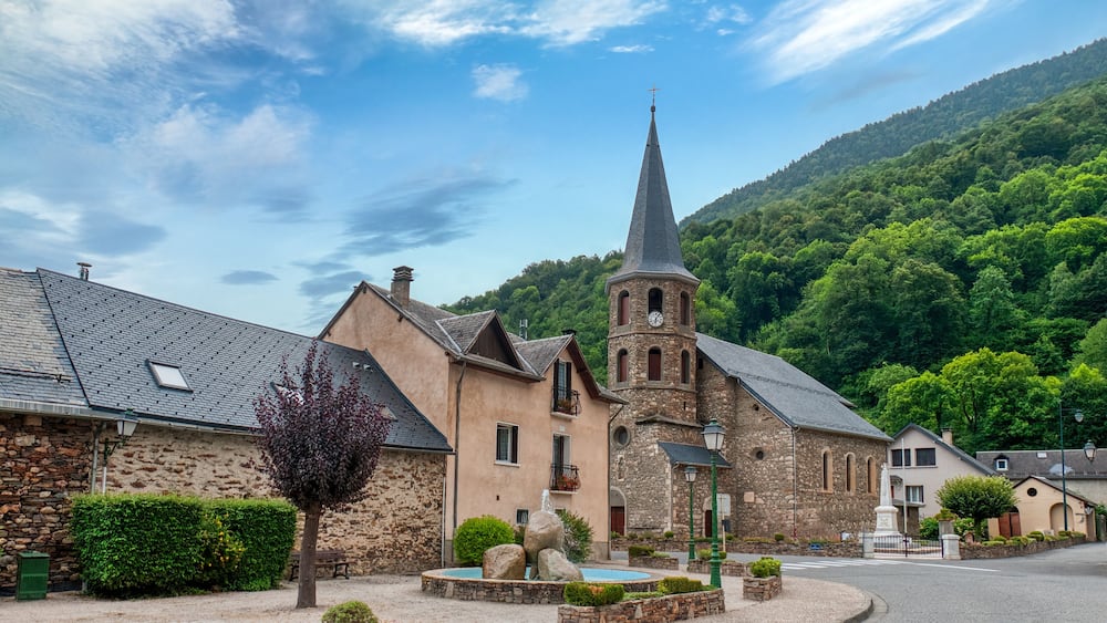 Une église avec un clocher imposant, un bâtiment en pierre et un arbre, le tout situé dans un cadre villageois.