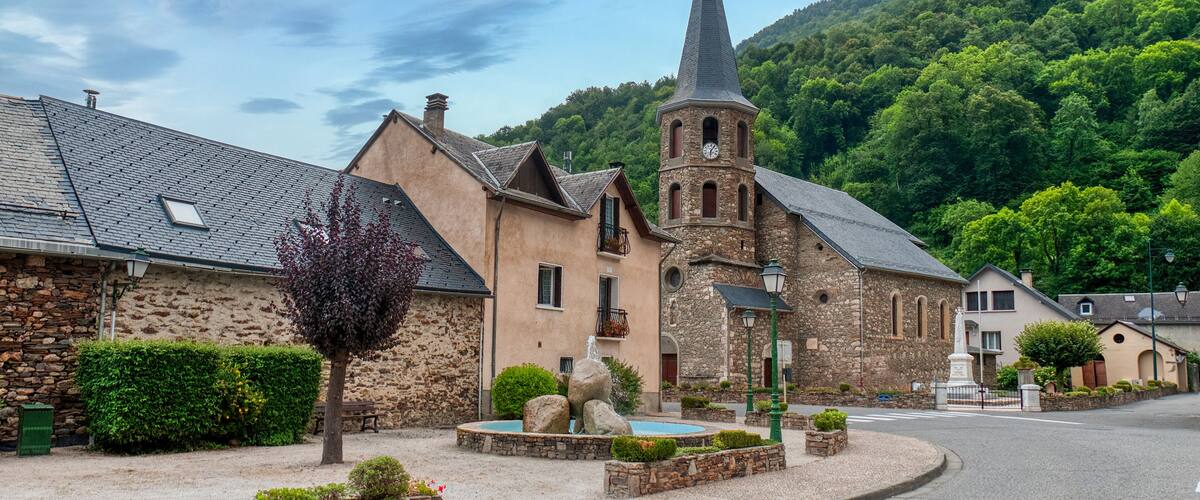 A church with a tall steeple, a stone building, and a tree in a village setting.