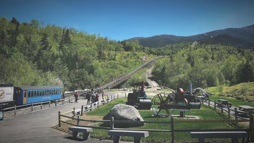 Un tren azul sobre rieles, un puente de madera, mesas de picnic y un paisaje montañoso.
