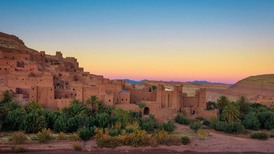 A desert village with adobe buildings, palm trees, and mountains in the background.
