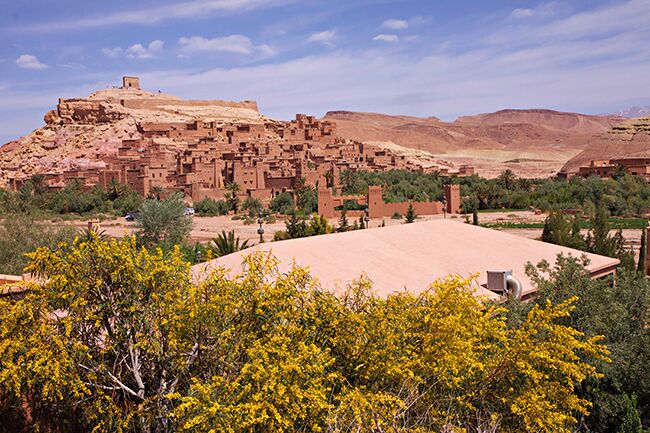 A desert landscape with a prominent red-brick structure on a hill, surrounded by sparse vegetation and distant mountains.