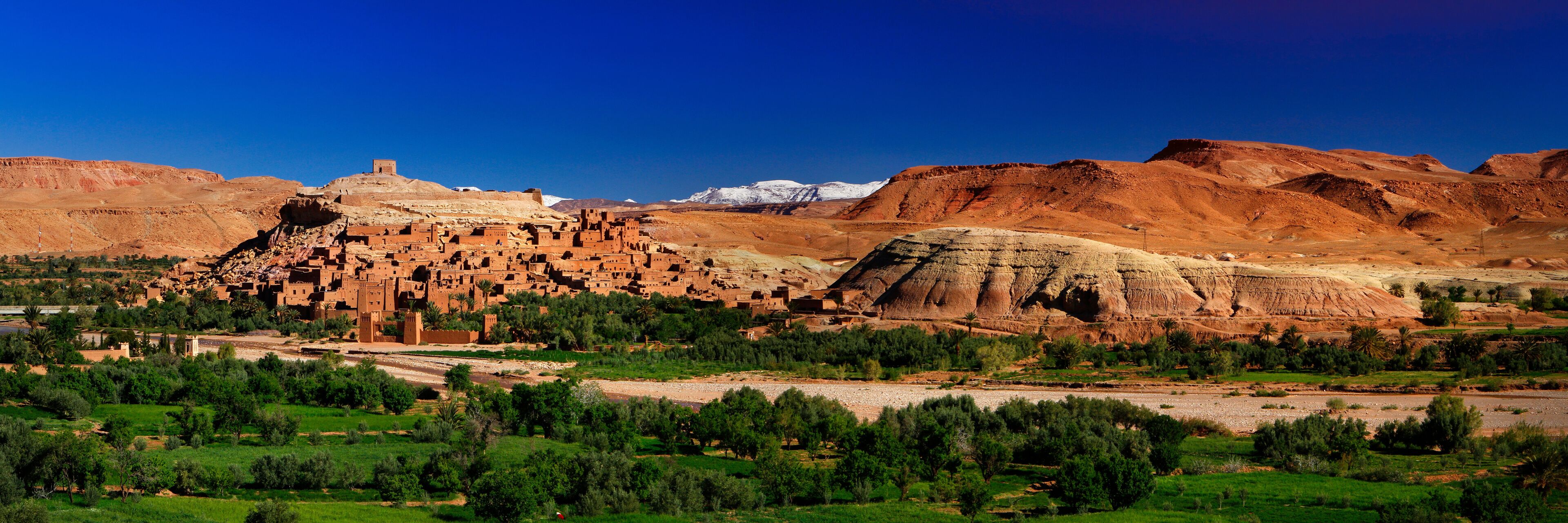 A desert landscape with a traditional mud-brick village, green fields, and distant snow-capped mountains.