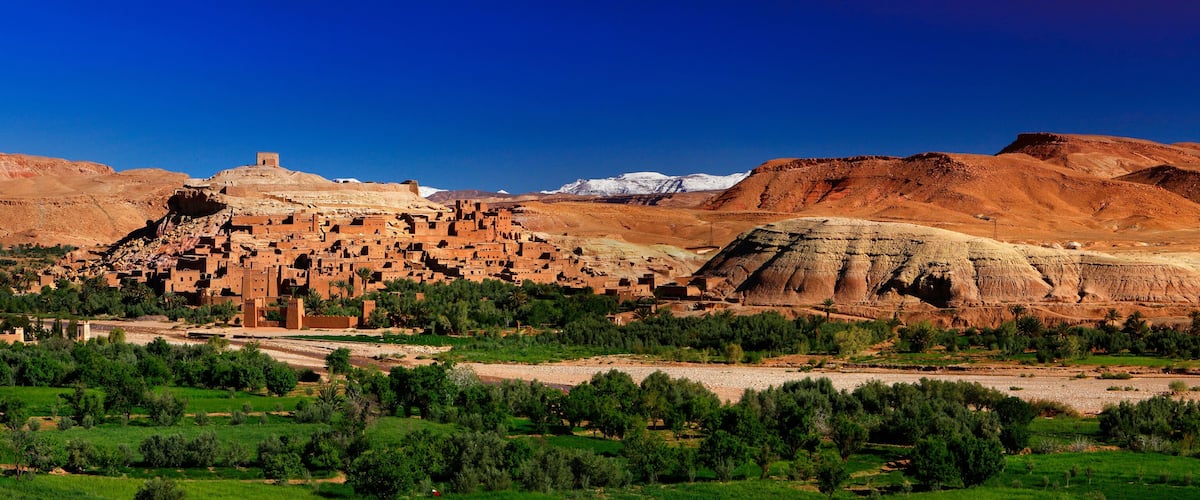 A desert landscape with a traditional mud-brick village, green fields, and distant snow-capped mountains.