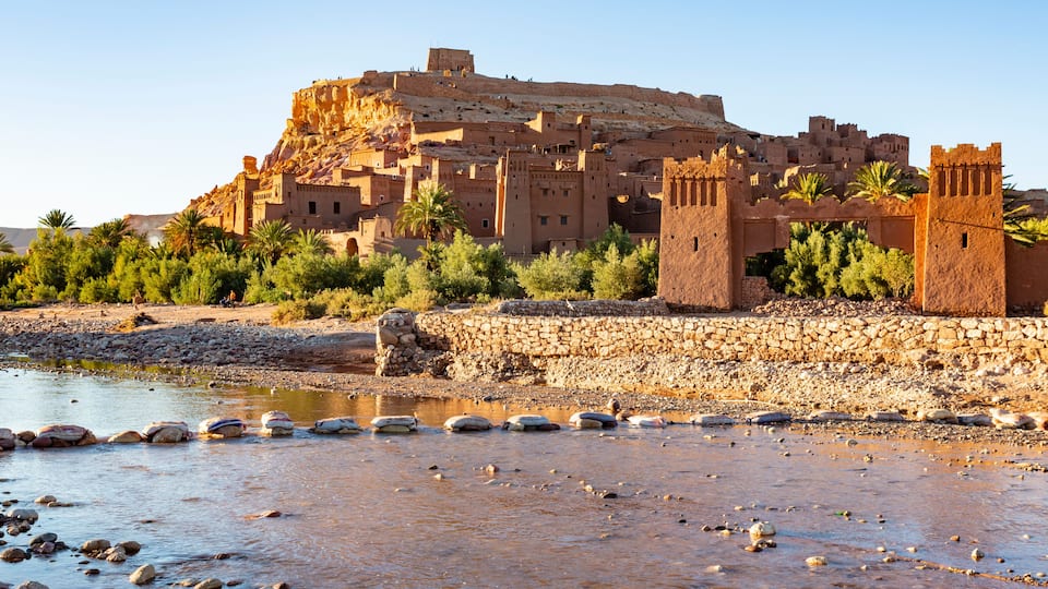 A fortified city with a prominent castle on a hill, palm trees, and a riverbank.