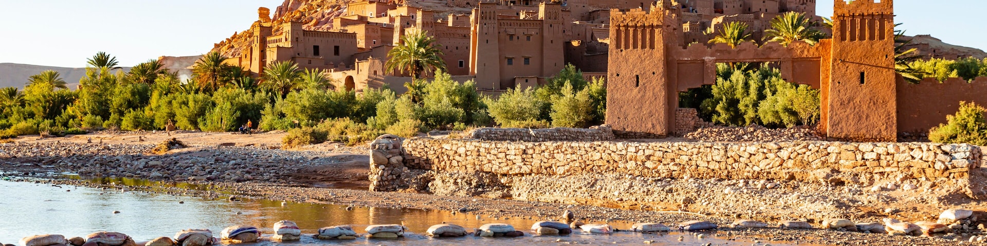 Una ciudad amurallada con un castillo imponente en una colina, palmeras y la orilla de un río.