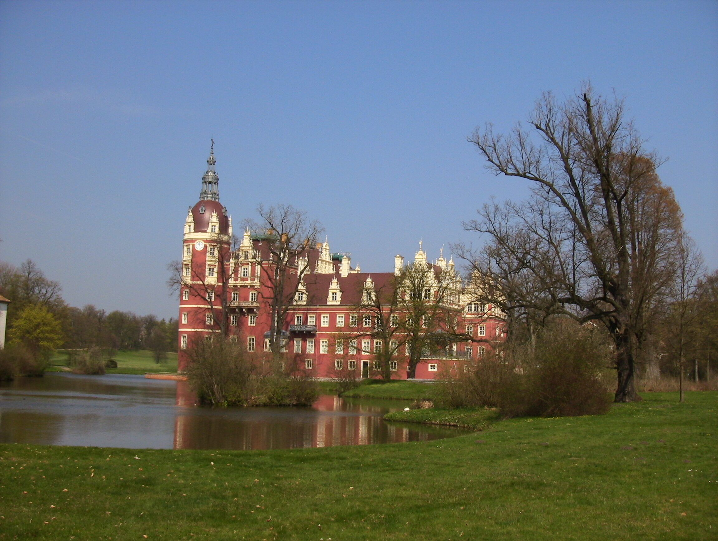 A large, ornate red building with a clock tower, surrounded by a lake and trees.