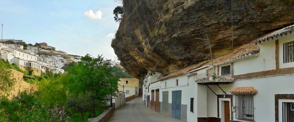 A narrow street flanked by white buildings with terracotta roofs, under a large rock formation.