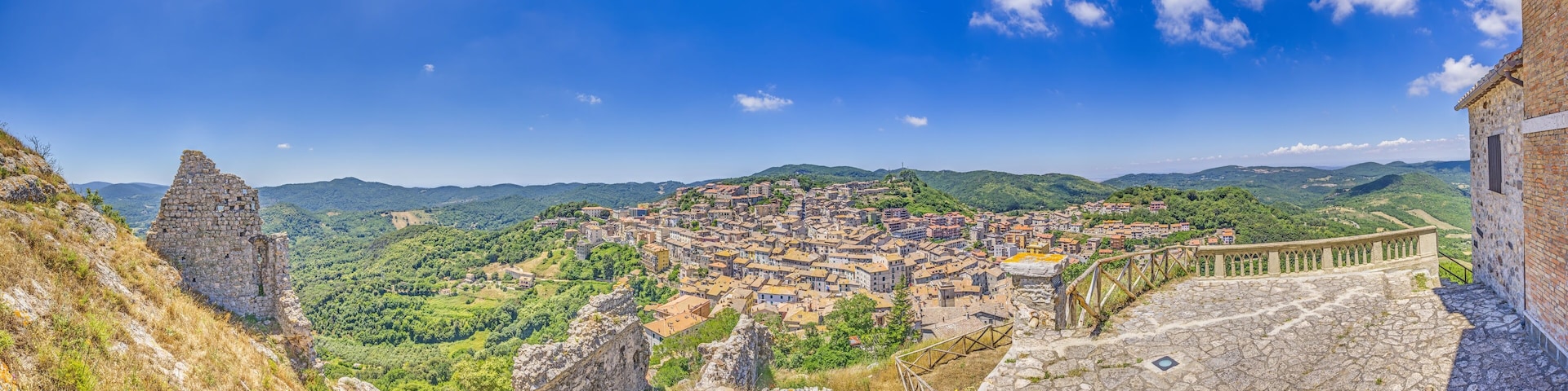 Vista panorámica de un pueblo histórico enclavado en un valle, con un muro de piedra y ruinas en primer plano.