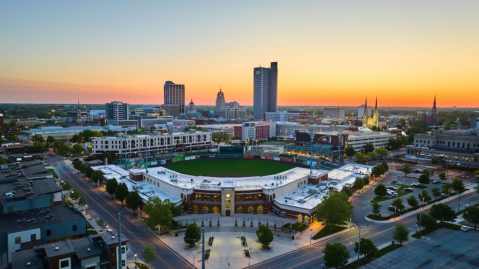 A cityscape with a baseball stadium, downtown buildings, and a church spire at sunset.
