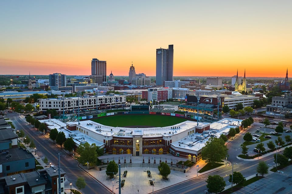 Paisagem urbana com um estádio de beisebol, edifícios do centro e a torre de uma igreja ao pôr do sol.