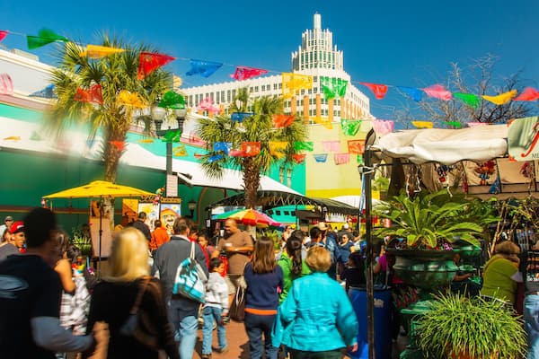 Um mercado ao ar livre movimentado, com bandeiras coloridas, palmeiras e um edifício imponente ao fundo.