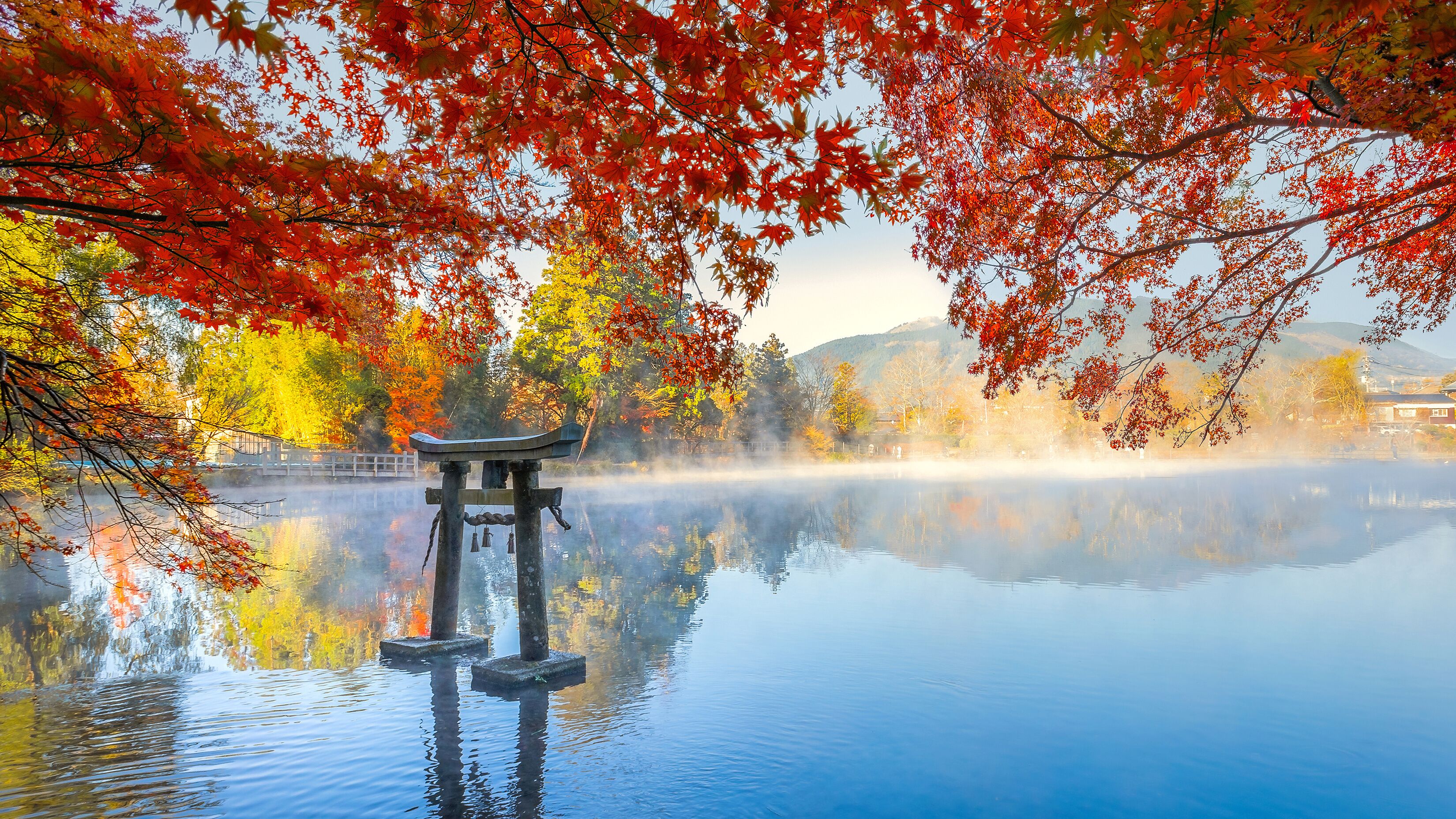 A serene lake with a traditional Japanese torii gate in the foreground, surrounded by autumn foliage and a misty mountain backdrop.