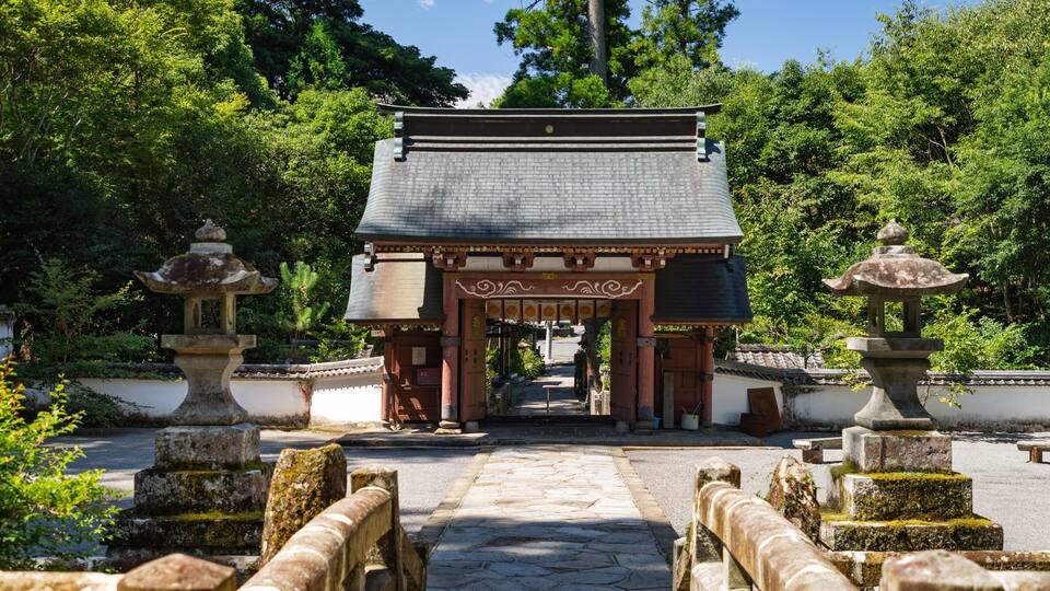 A traditional Japanese gate with a tiled roof, flanked by stone lanterns and surrounded by lush greenery.