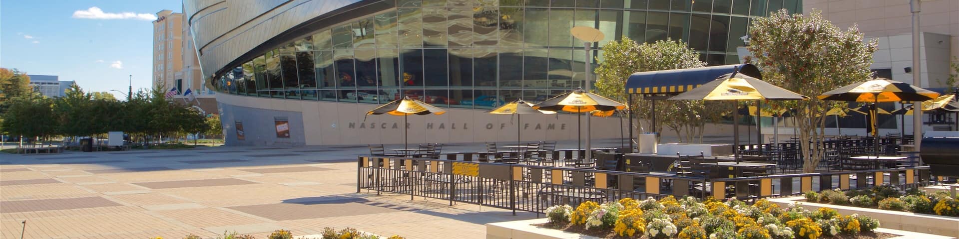 A modern building with a curved glass facade, outdoor seating with umbrellas, and a flower garden in front.