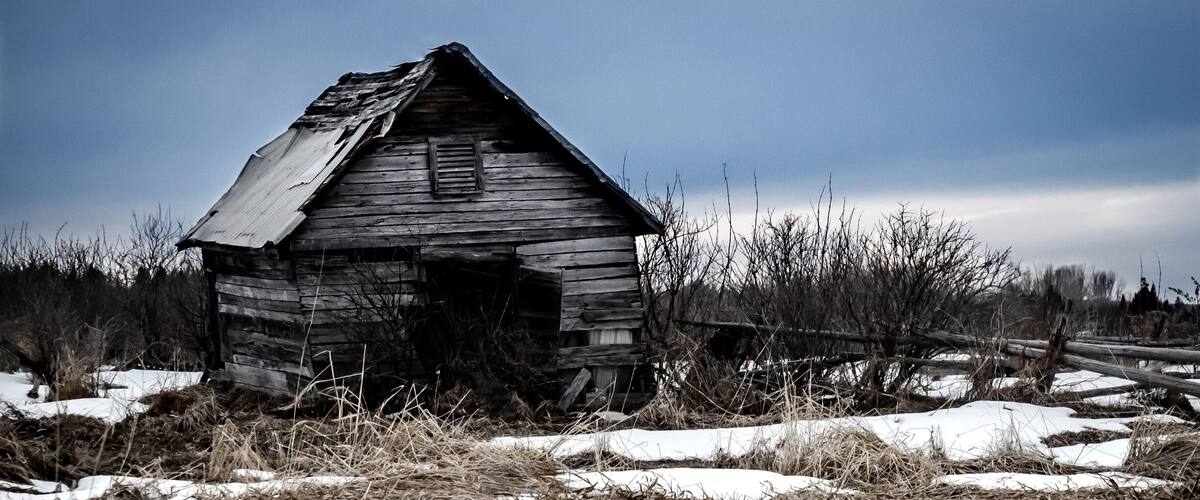 An old wooden cabin in a snowy field with overgrown grass and a fence in the background.