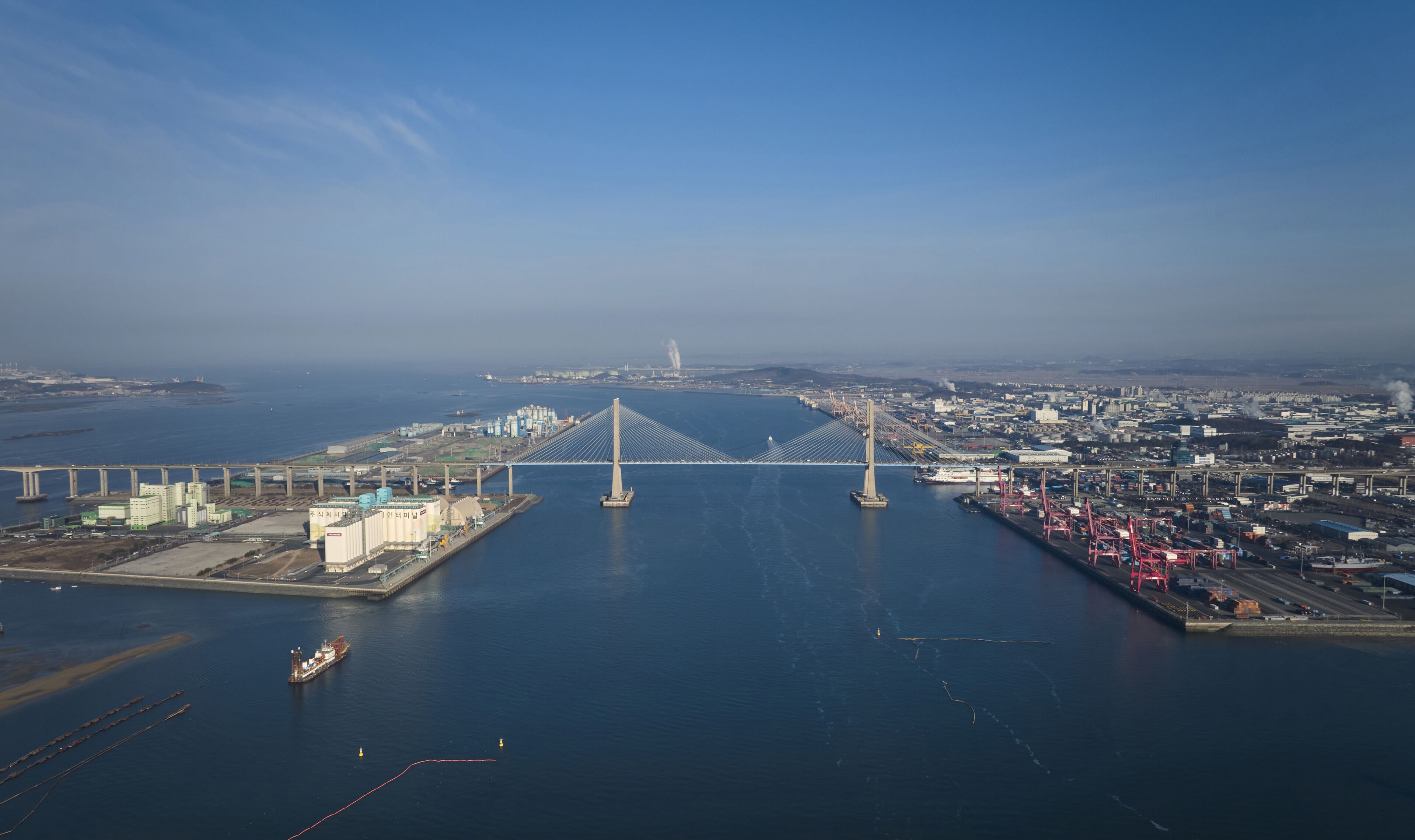 A large suspension bridge spanning a wide river, with industrial buildings and a port on either side.