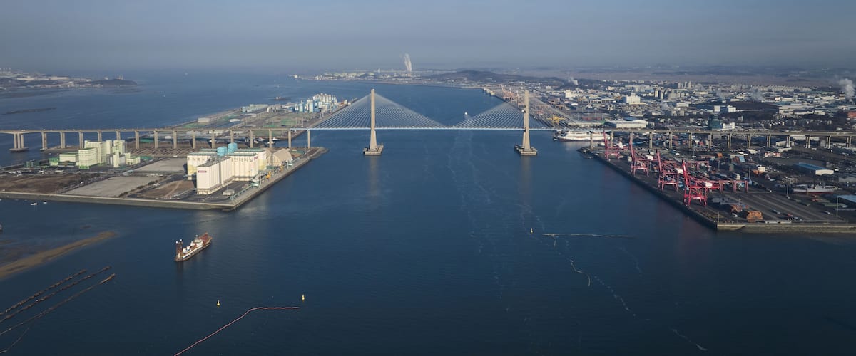 A large suspension bridge spanning a wide river, with industrial buildings and a port on either side.
