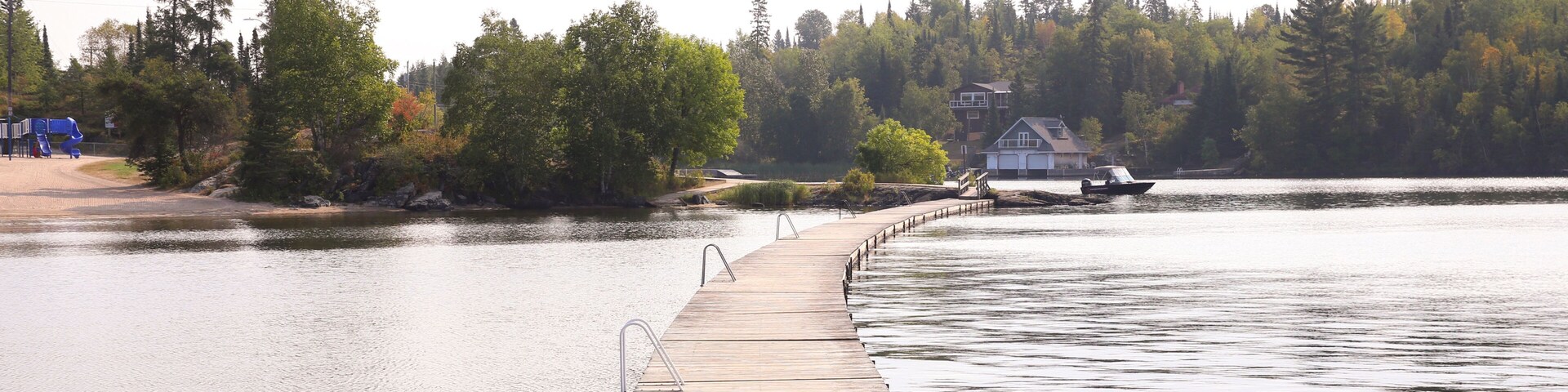 Un muelle de madera se adentra en un lago tranquilo, rodeado de un bosque denso y una casa en la orilla opuesta.