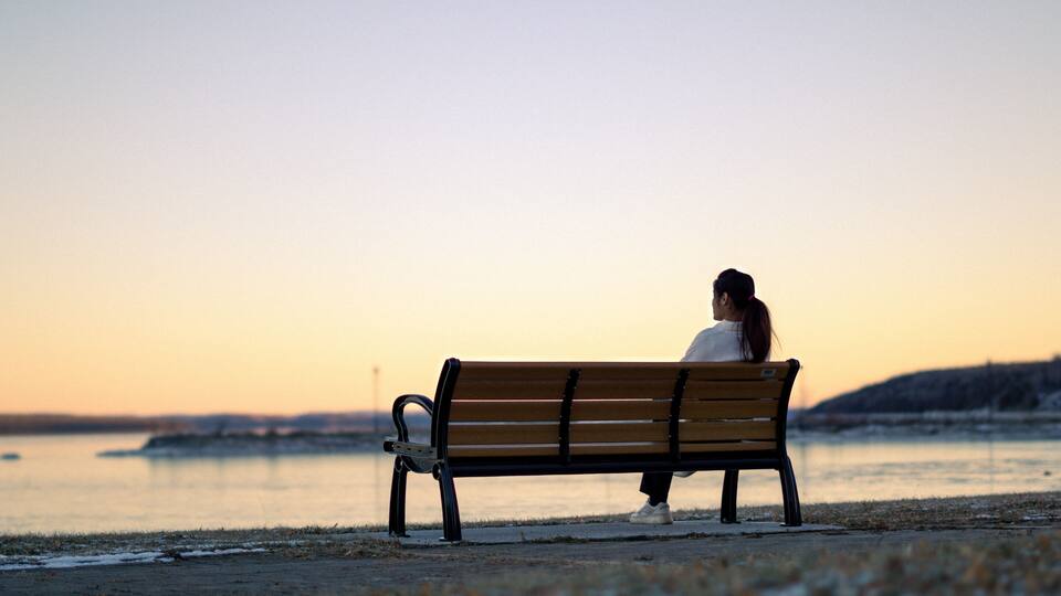 A woman sitting on a bench by a body of water during sunset.