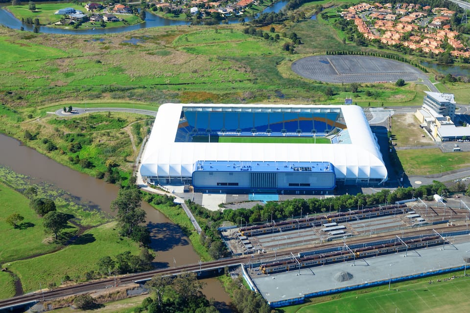 An aerial view of a stadium with a large roof, surrounded by green fields, a river, and a train station.