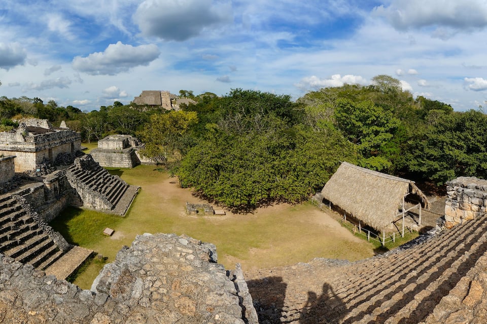 Ancient stone ruins with stepped platforms and a thatched-roof structure amidst lush greenery.