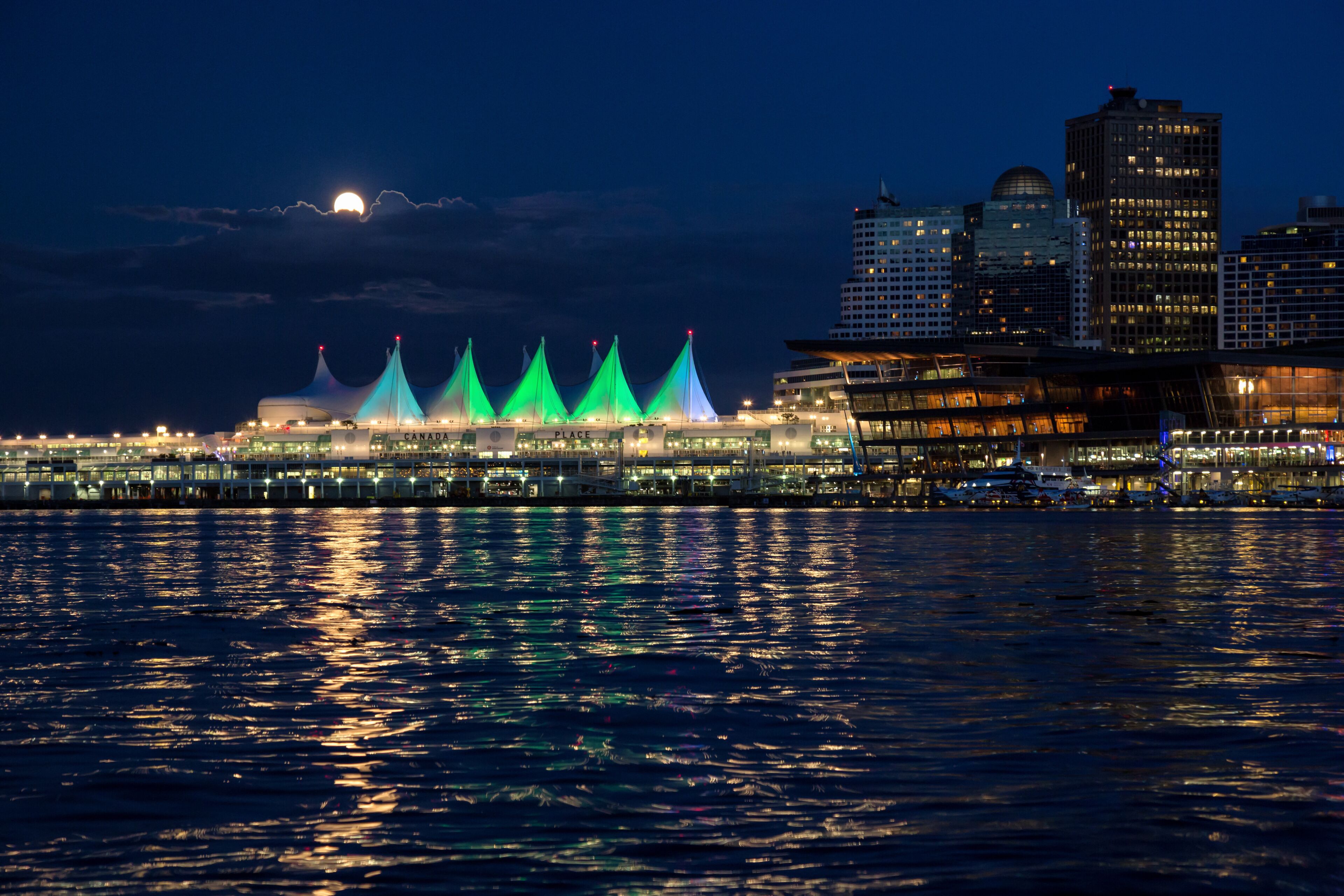 A waterfront cityscape with illuminated buildings and a prominent structure with green lights reflecting on the water.