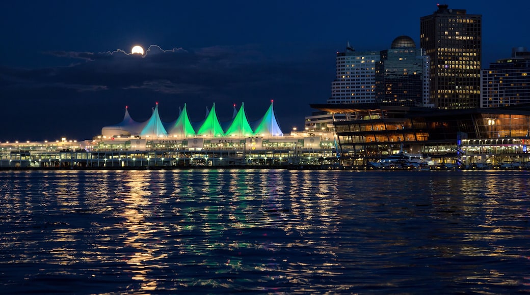 A waterfront cityscape with illuminated buildings and a prominent structure with green lights reflecting on the water.