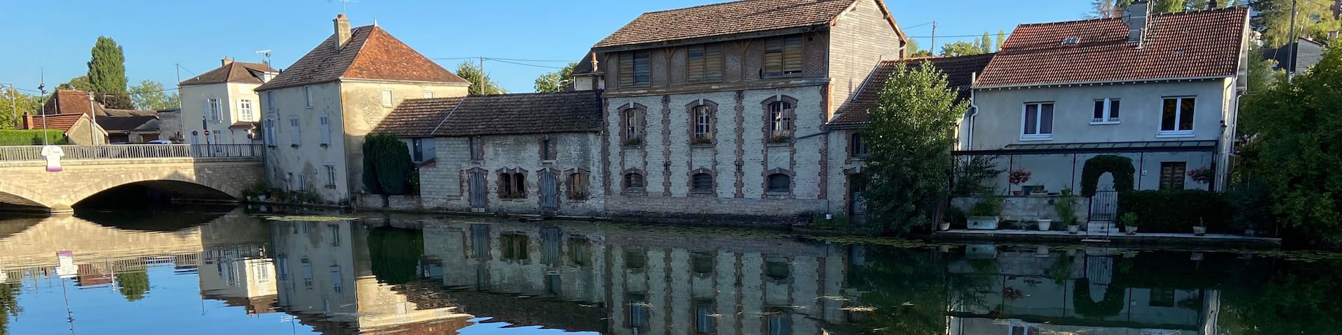 A calm river reflecting buildings, a stone bridge, and lily pads.